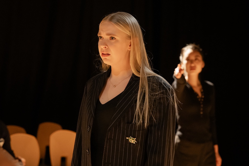 Actor stands with a distraught expression on her face while another actor stands behind her, looking on.